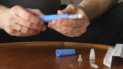 A person’s hands assemble a blue injectable pen device on a wooden table, with a cap, needle tip, and packaging laid out nearby.