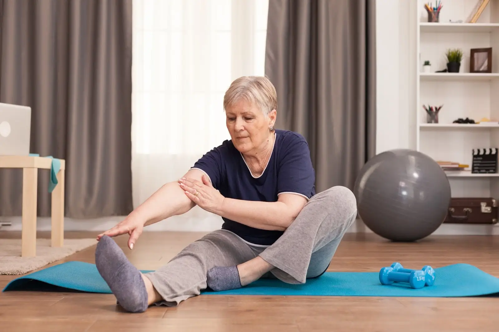 An older woman sits on a yoga mat at home, stretching to touch her foot with one hand and touching her leg with the other. Exercise equipment and a stability ball are visible in the background.