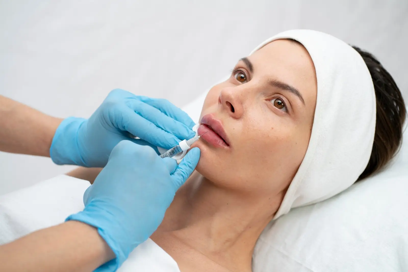 A woman wearing a white headband receives a cosmetic injection in her lips from a professional wearing blue gloves. She is lying on a treatment bed in a clinical setting.