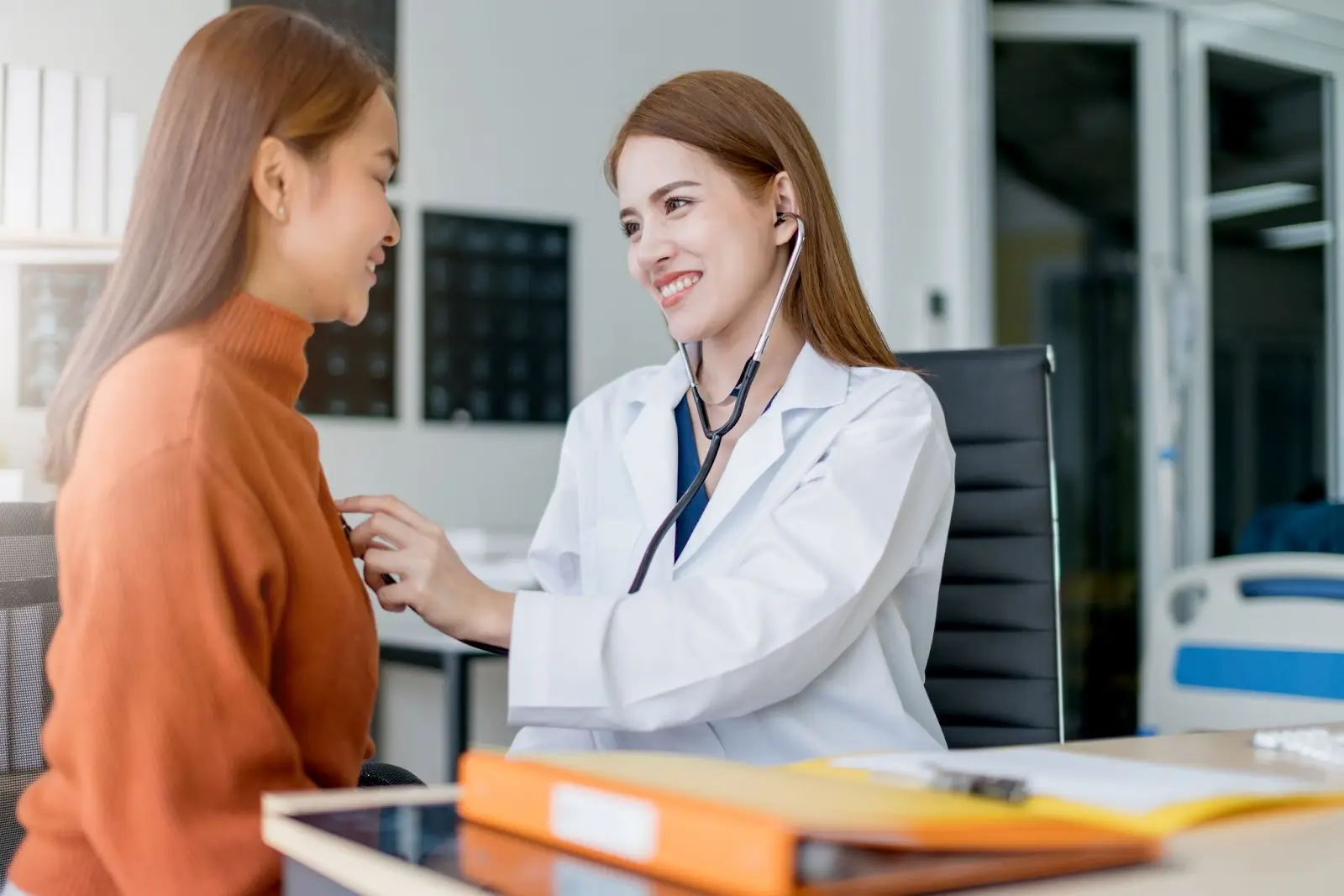 A female doctor, smiling, uses a stethoscope to examine a young woman in an orange sweater during a consultation in a bright medical office.