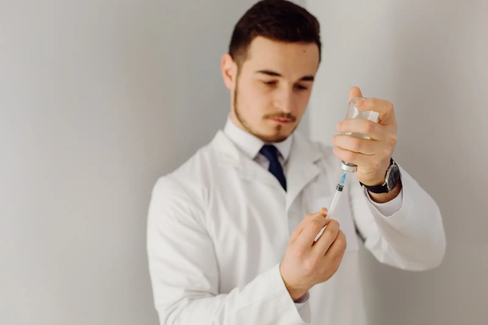 A doctor in a white coat prepares a syringe by drawing liquid from a vial, focusing intently on the task against a plain background.