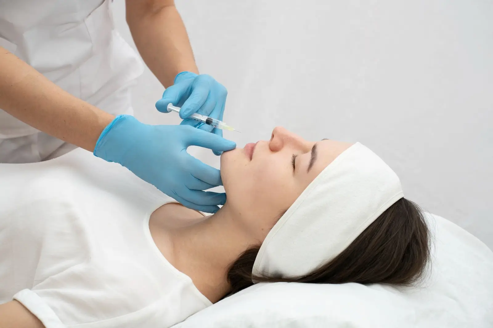 A woman lies on a treatment bed with her eyes closed while a medical professional wearing blue gloves injects her chin. The woman wears a white headband and shirt. The setting appears clinical and clean.