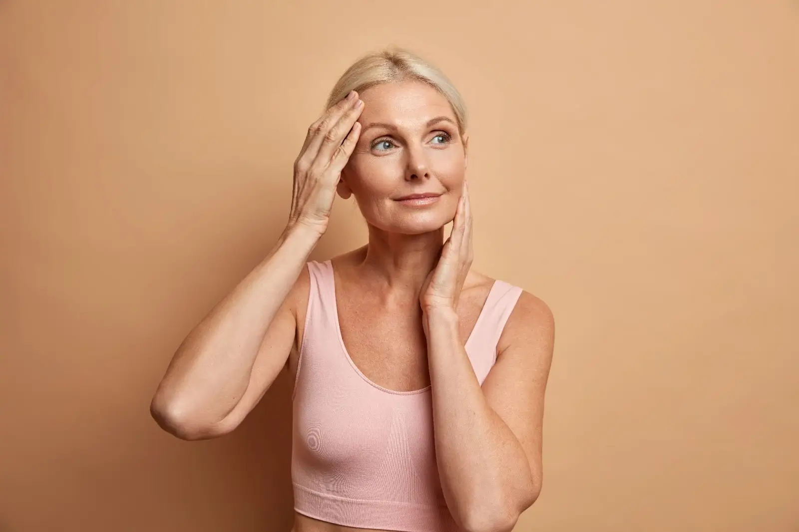 A middle-aged woman with blonde hair wearing a light pink tank top stands against a beige background, touching her face with both hands and looking slightly to the side with a gentle expression.