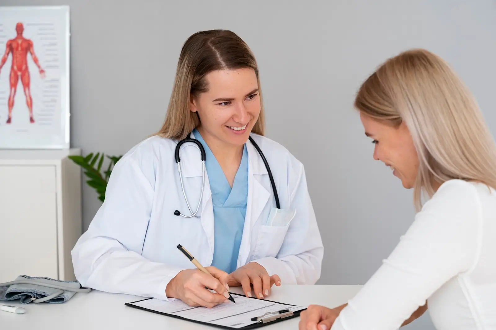 A female doctor with a stethoscope smiles while talking to a patient and writing on a clipboard at a desk in a medical office. A medical anatomy poster is visible in the background.