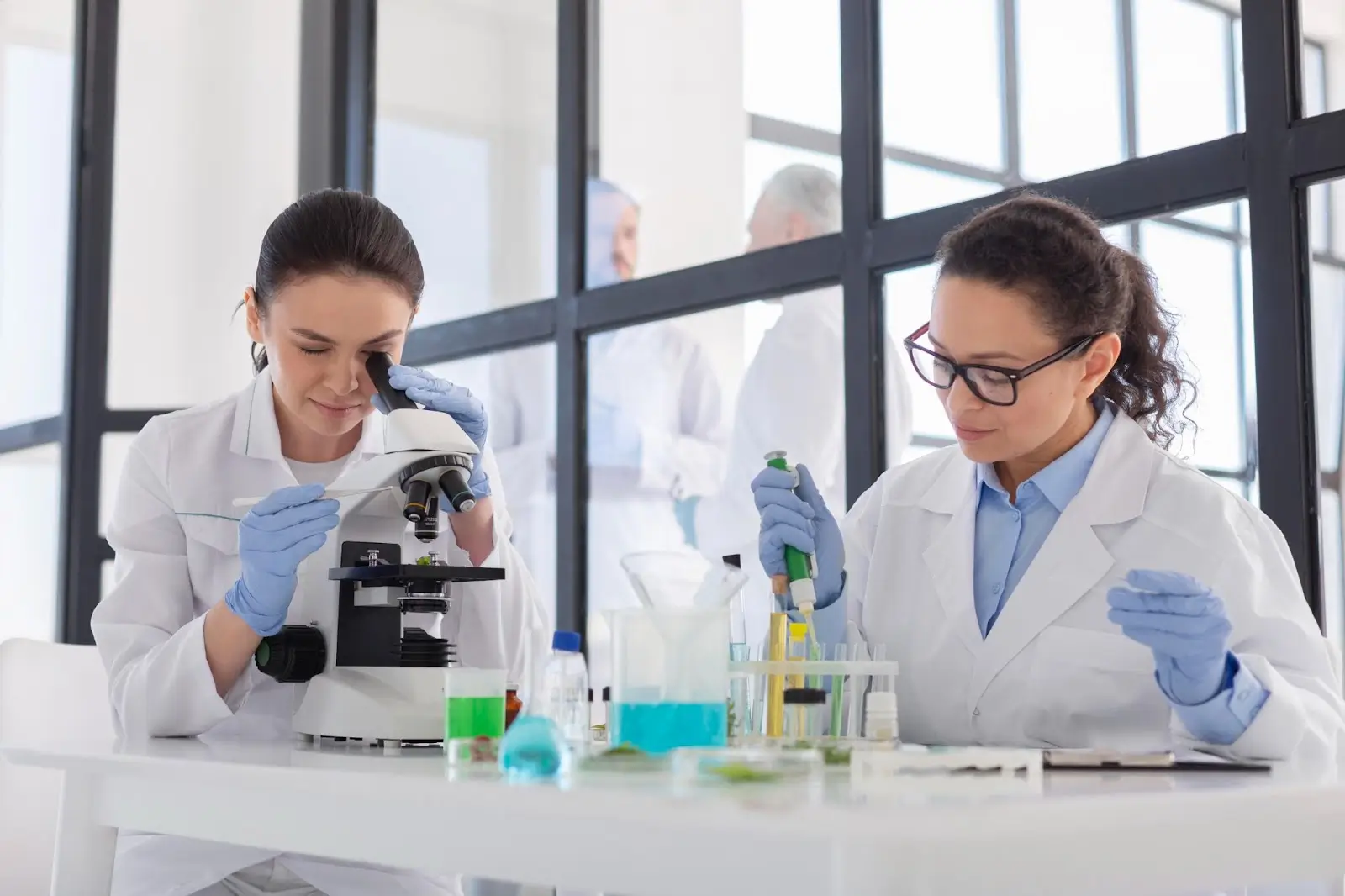 Two female scientists in lab coats work in a laboratory; one looks into a microscope, while the other uses a pipette with test tubes containing colorful liquids. Lab equipment is visible on the table.