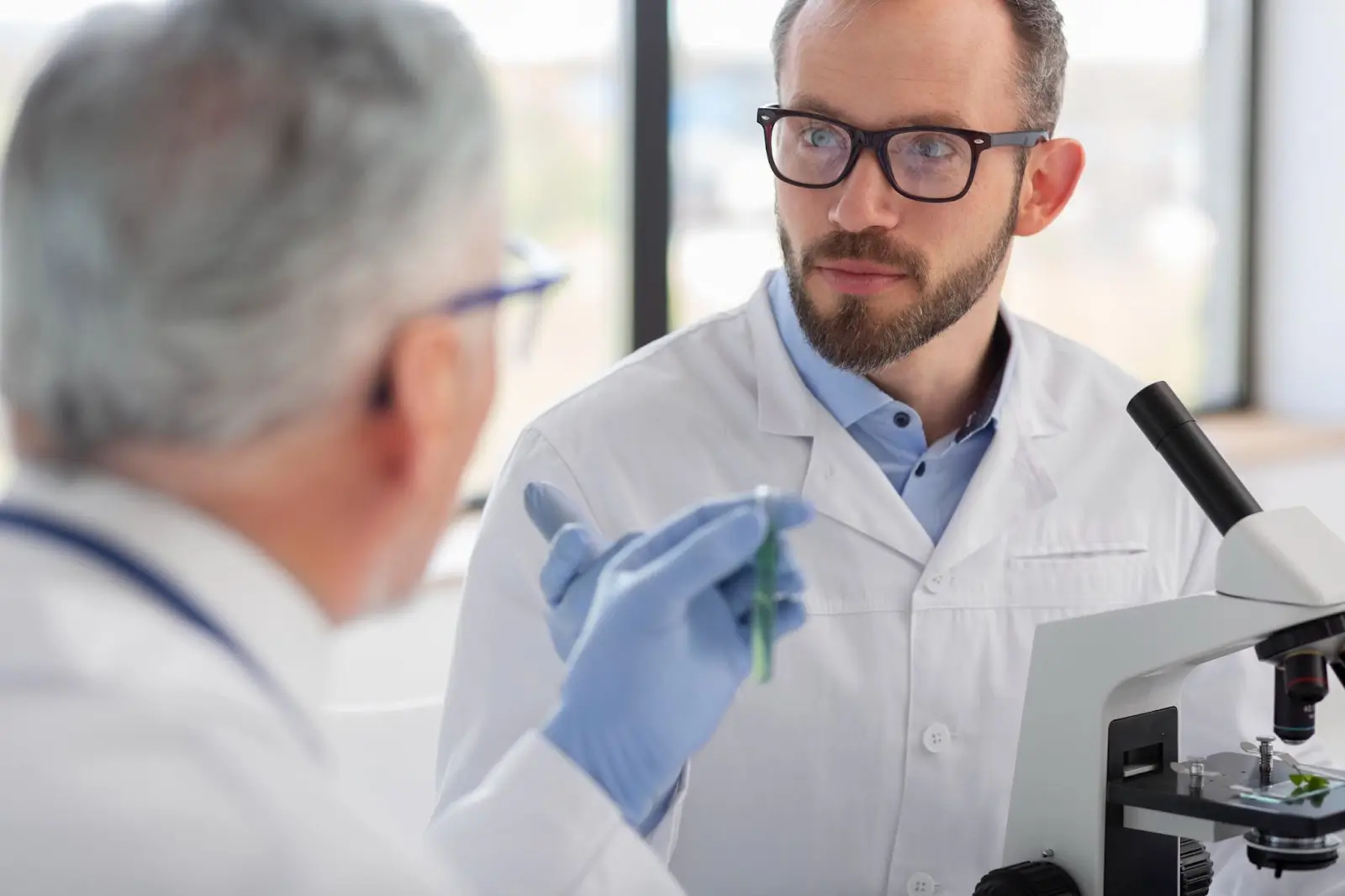 Two male scientists in lab coats have a discussion by a microscope. One, with blue gloves, holds a test tube while the other listens attentively. Bright laboratory setting with large windows in the background.