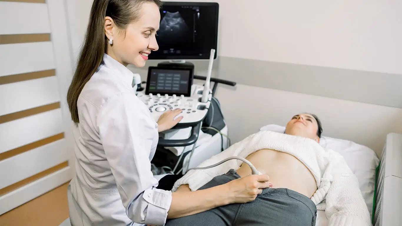 A smiling medical professional performs an ultrasound scan on a woman lying on an examination table, with an ultrasound machine and monitor in the background.