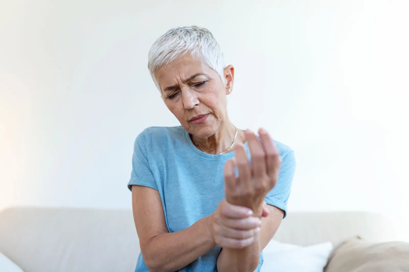 Older woman with short gray hair wearing a blue shirt sits on a couch, holding her wrist and looking at it with a pained expression, suggesting discomfort or pain in her wrist.