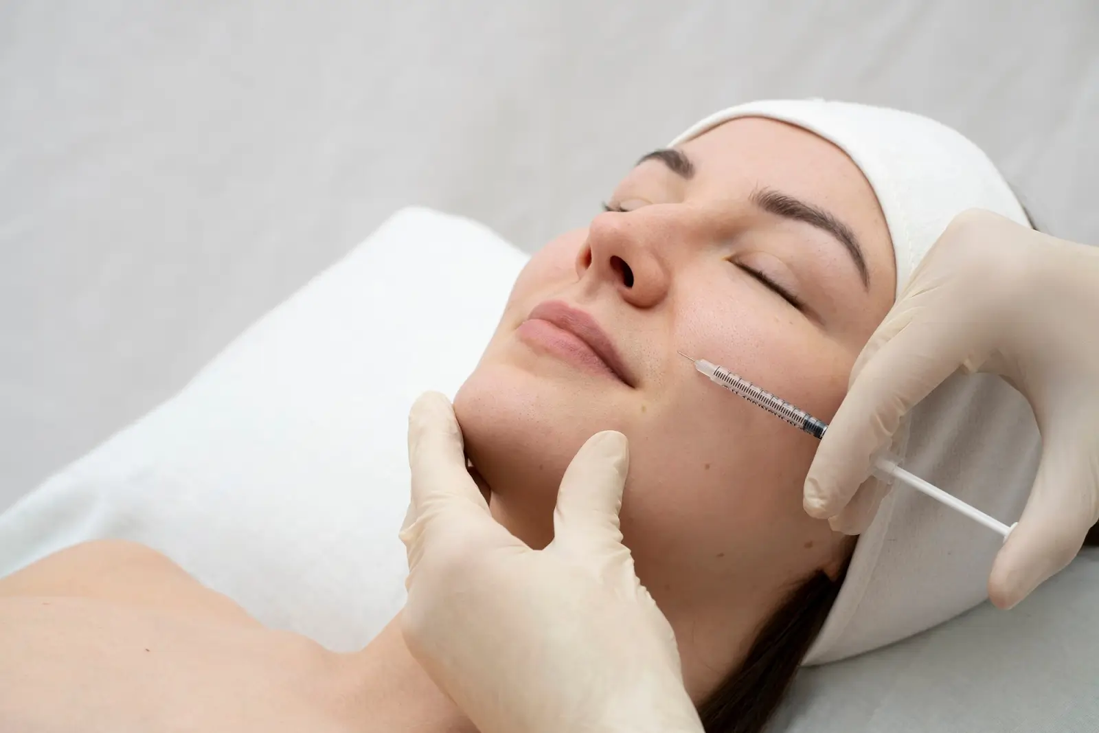A woman lying on a treatment table with her eyes closed is receiving an injection in her cheek. She is wearing a white headband, and the person administering the injection is wearing white gloves.