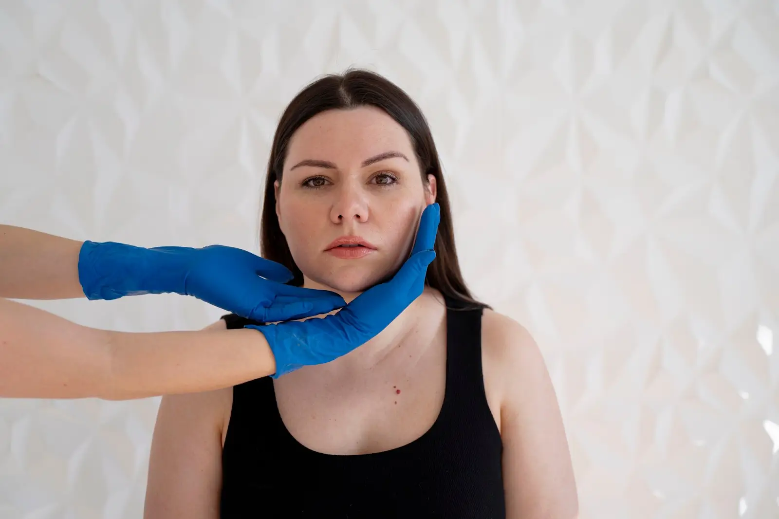 A woman with straight dark hair and a neutral expression sits facing the camera while two gloved hands gently touch her chin and cheek, against a textured white background.