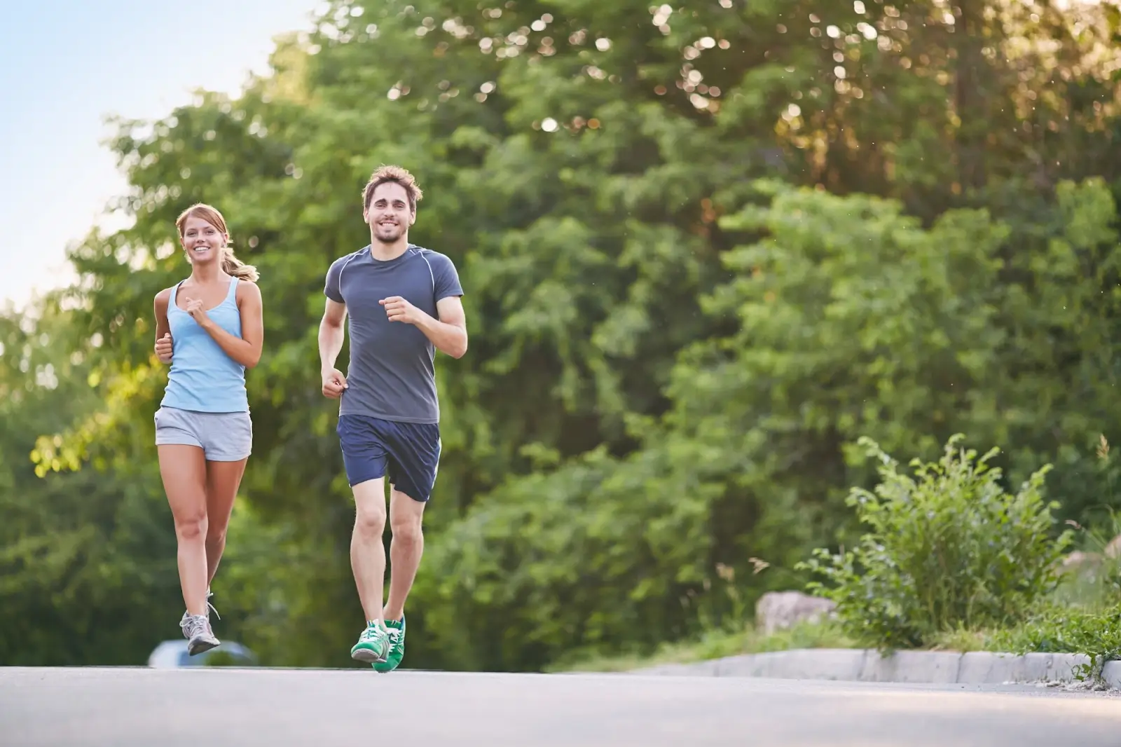 A man and a woman in athletic clothing are jogging together on a paved path outdoors, surrounded by lush green trees on a sunny day. Both appear to be smiling and enjoying the activity.
