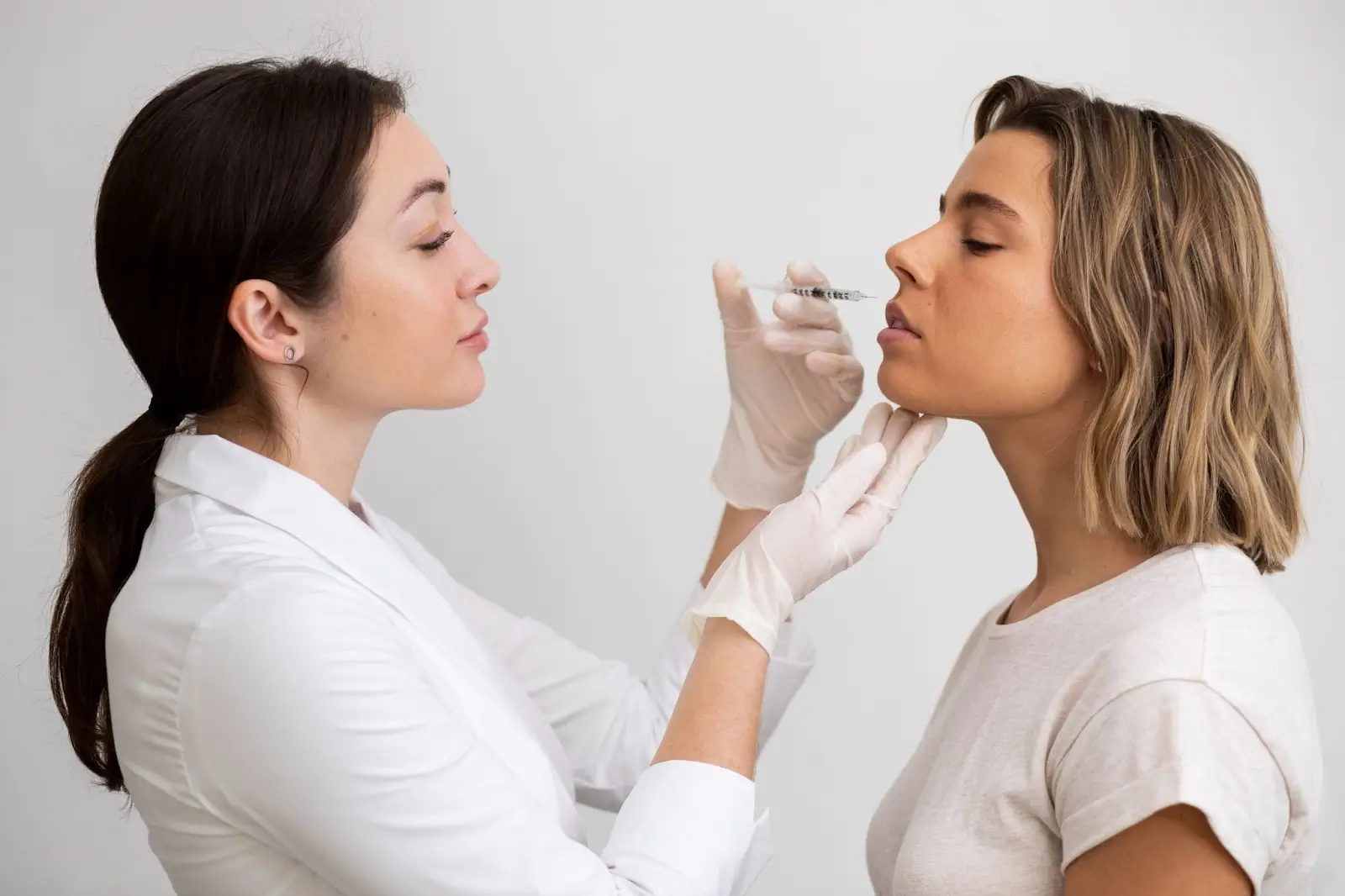 A female doctor wearing gloves prepares to inject a syringe into a young woman's face, who is sitting calmly and tilting her chin up, possibly for a cosmetic procedure.