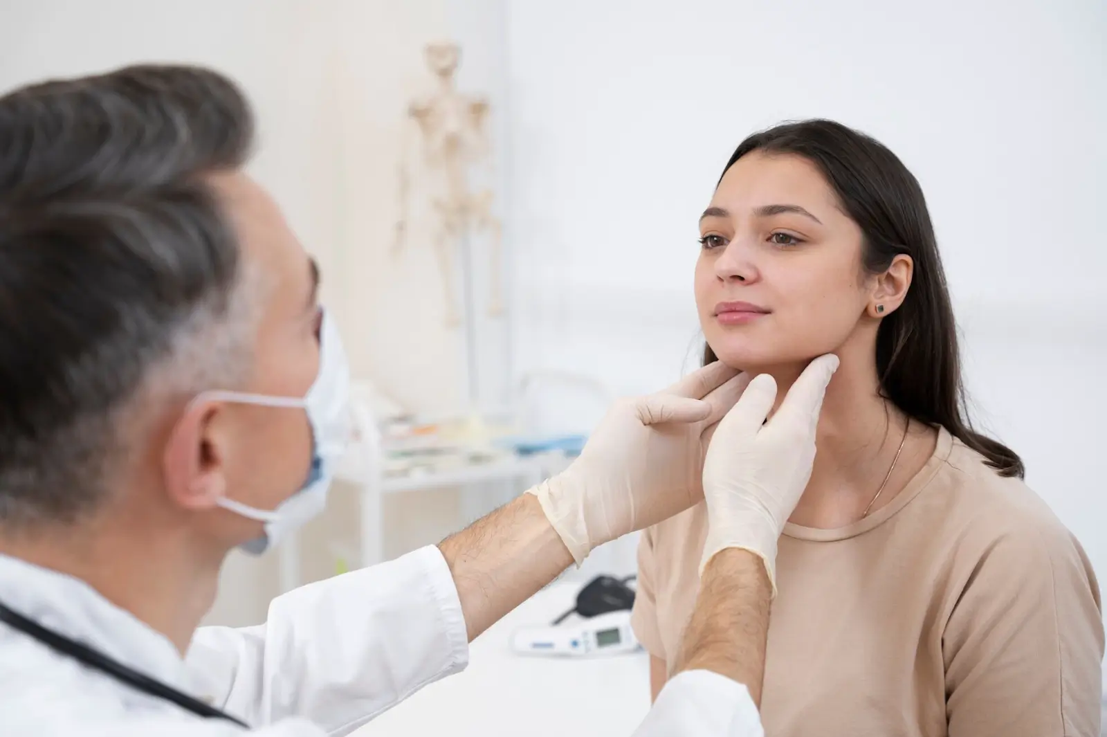 A doctor wearing gloves and a mask examines a young woman's neck in a medical office, possibly checking her lymph nodes or thyroid. The woman sits calmly, looking slightly upward.