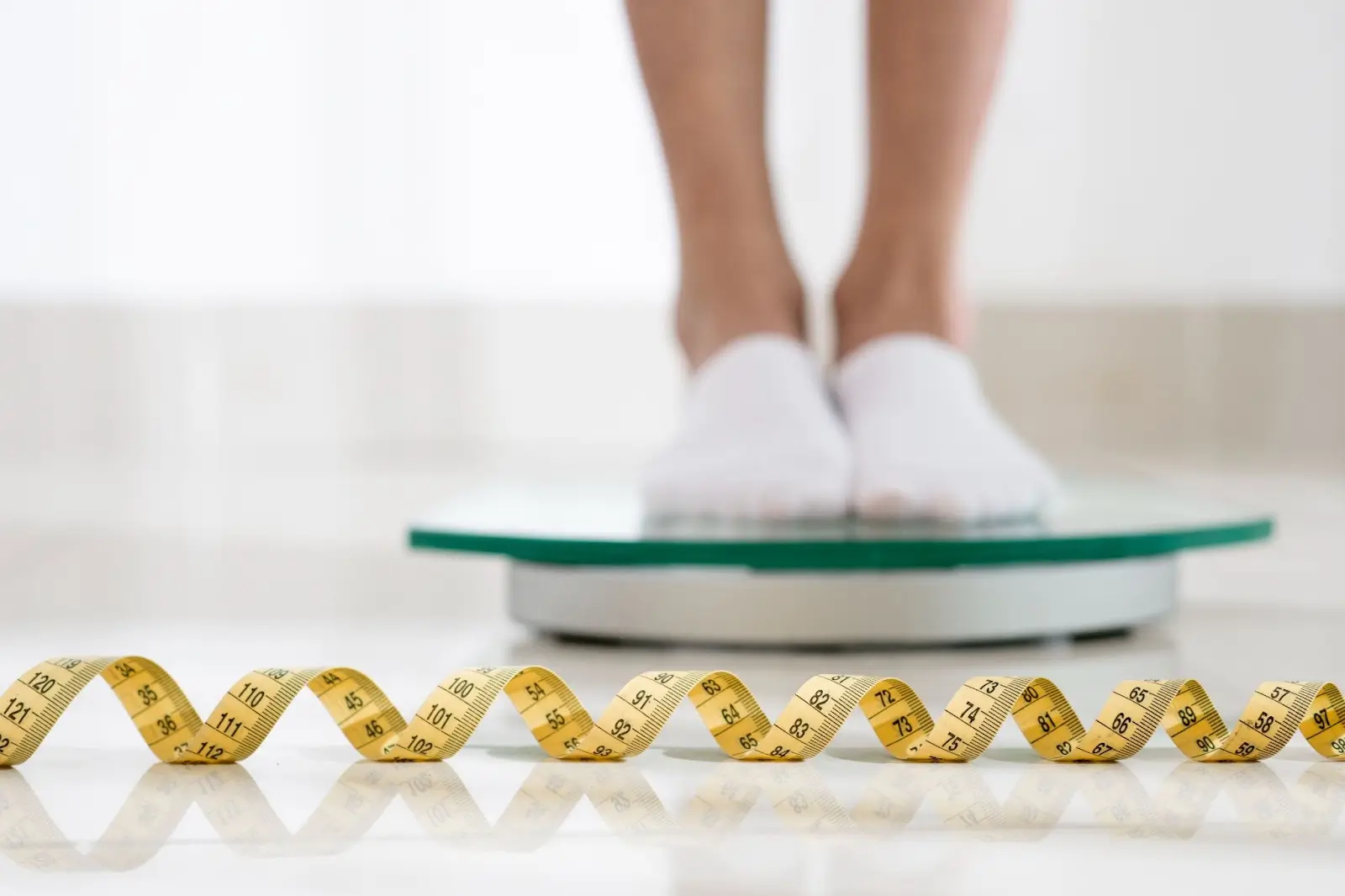 A person wearing white socks stands on a glass weighing scale, with a yellow measuring tape curled on the floor in the foreground.