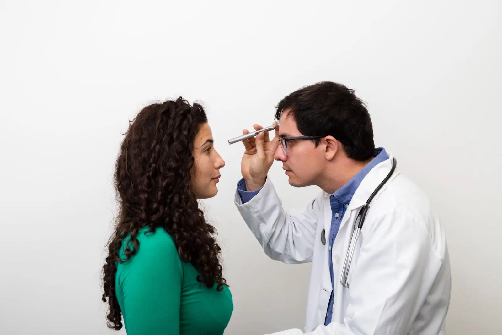 A doctor in a white coat holds a penlight and examines the eyes of a woman with curly hair wearing a green top, against a plain white background.
