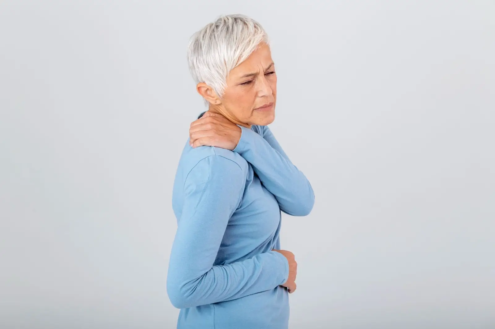 An older woman with short gray hair wearing a blue long-sleeve shirt holds her shoulder and appears to be in pain, with a pained expression on her face against a plain background.