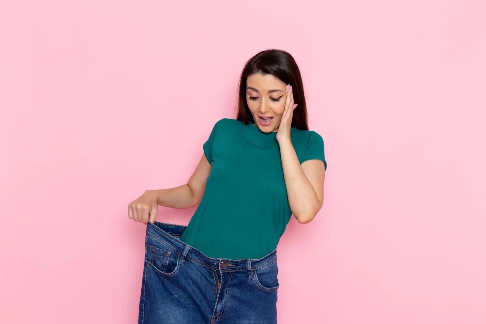 A surprised woman in a green shirt holds out the waistband of loose blue jeans, showing they are too large for her, while standing against a pink background.