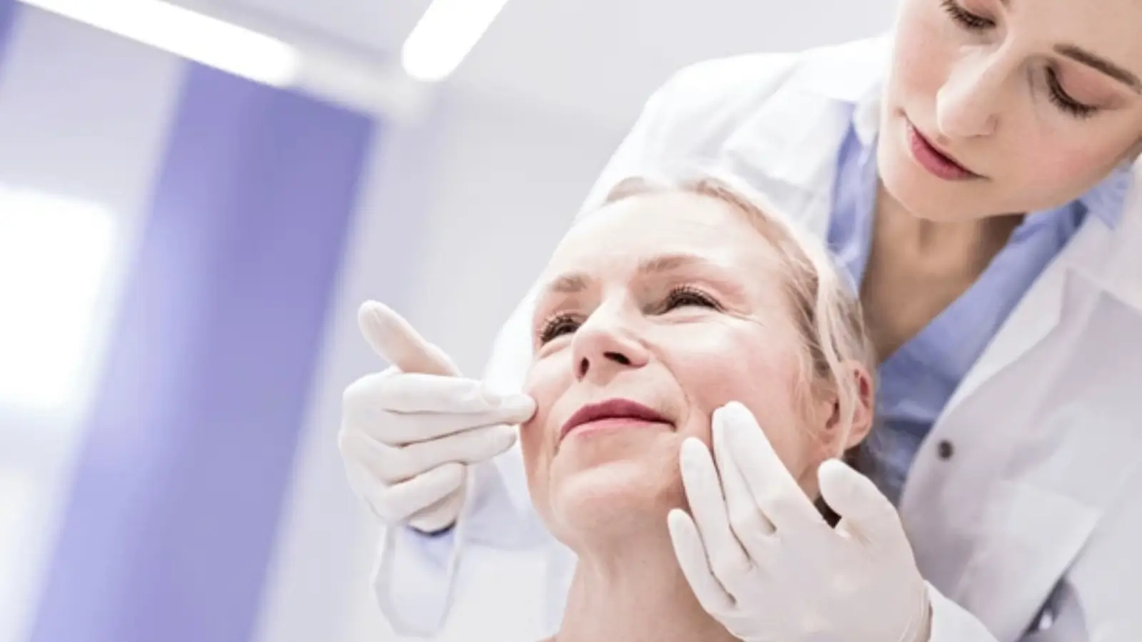 A doctor wearing white gloves examines the face of an older woman in a clinical setting. The doctor gently touches the woman's cheeks while the woman looks upward, appearing relaxed.