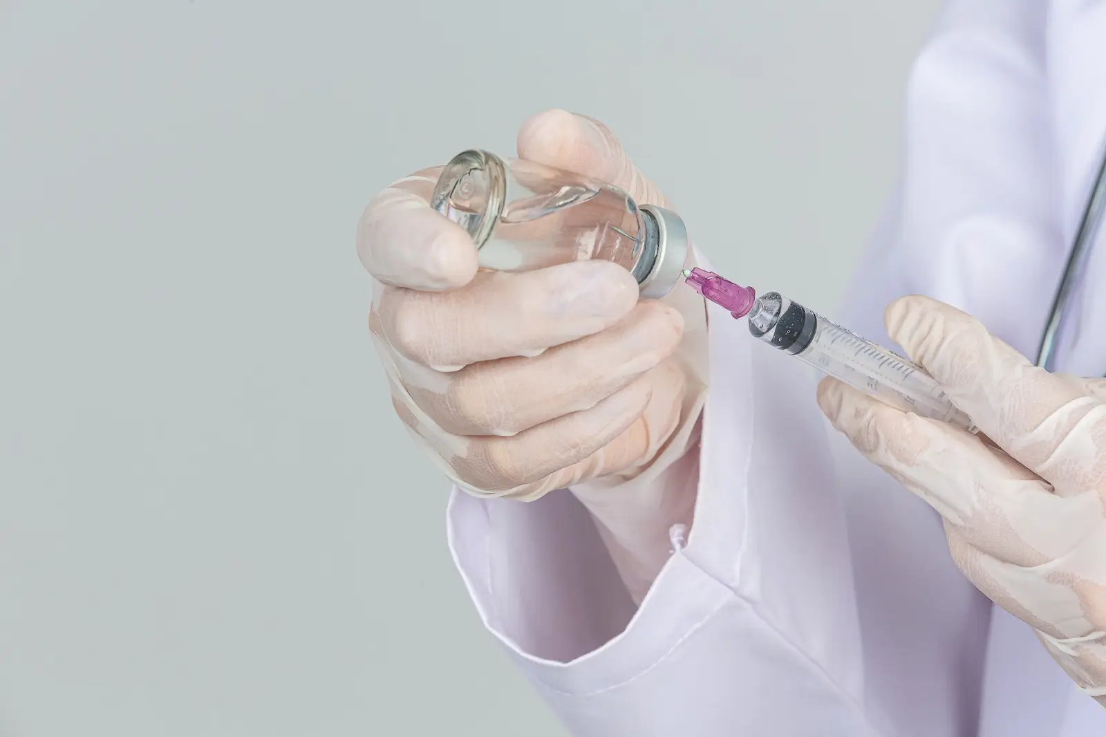 A person wearing white medical gloves and a lab coat draws liquid from a vial into a syringe, preparing for an injection.