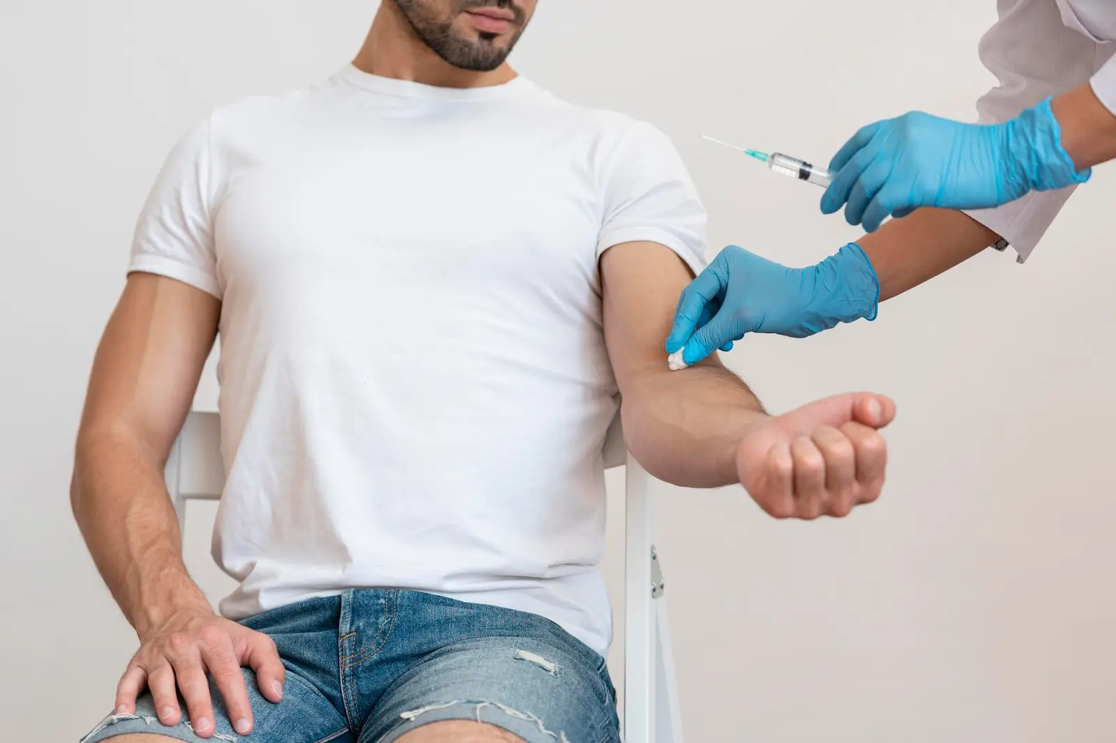 A person in a white t-shirt and denim shorts sits on a chair while a healthcare worker wearing blue gloves prepares to give an injection in their arm.