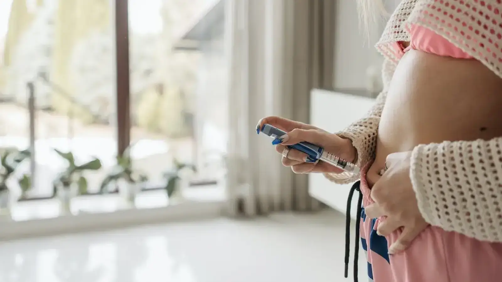A person in pink pajamas injects themselves in the abdomen with an insulin pen while standing indoors near a window with potted plants on the sill.