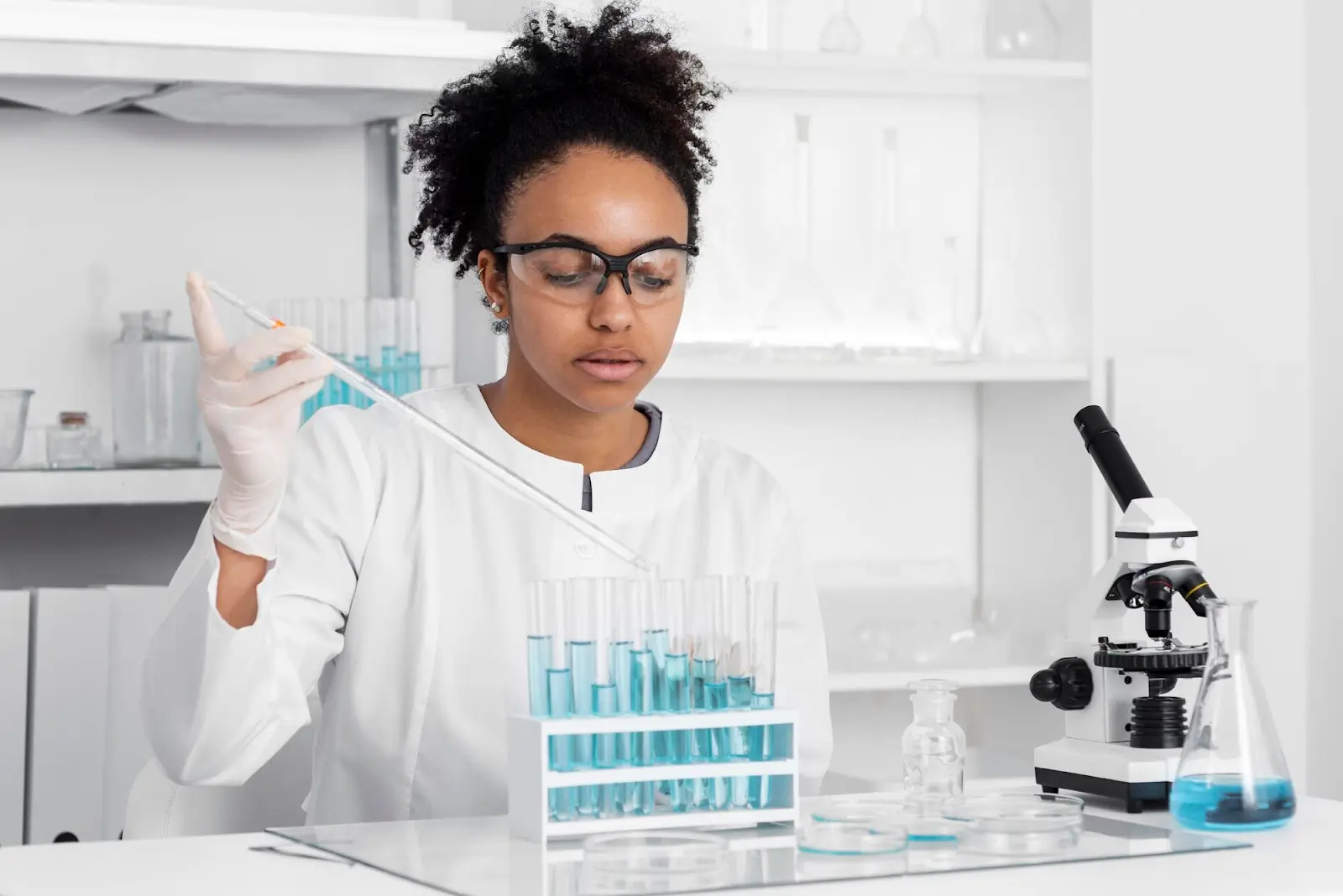 A scientist wearing safety goggles and gloves uses a pipette to transfer blue liquid into test tubes in a laboratory, with a microscope and glassware nearby.