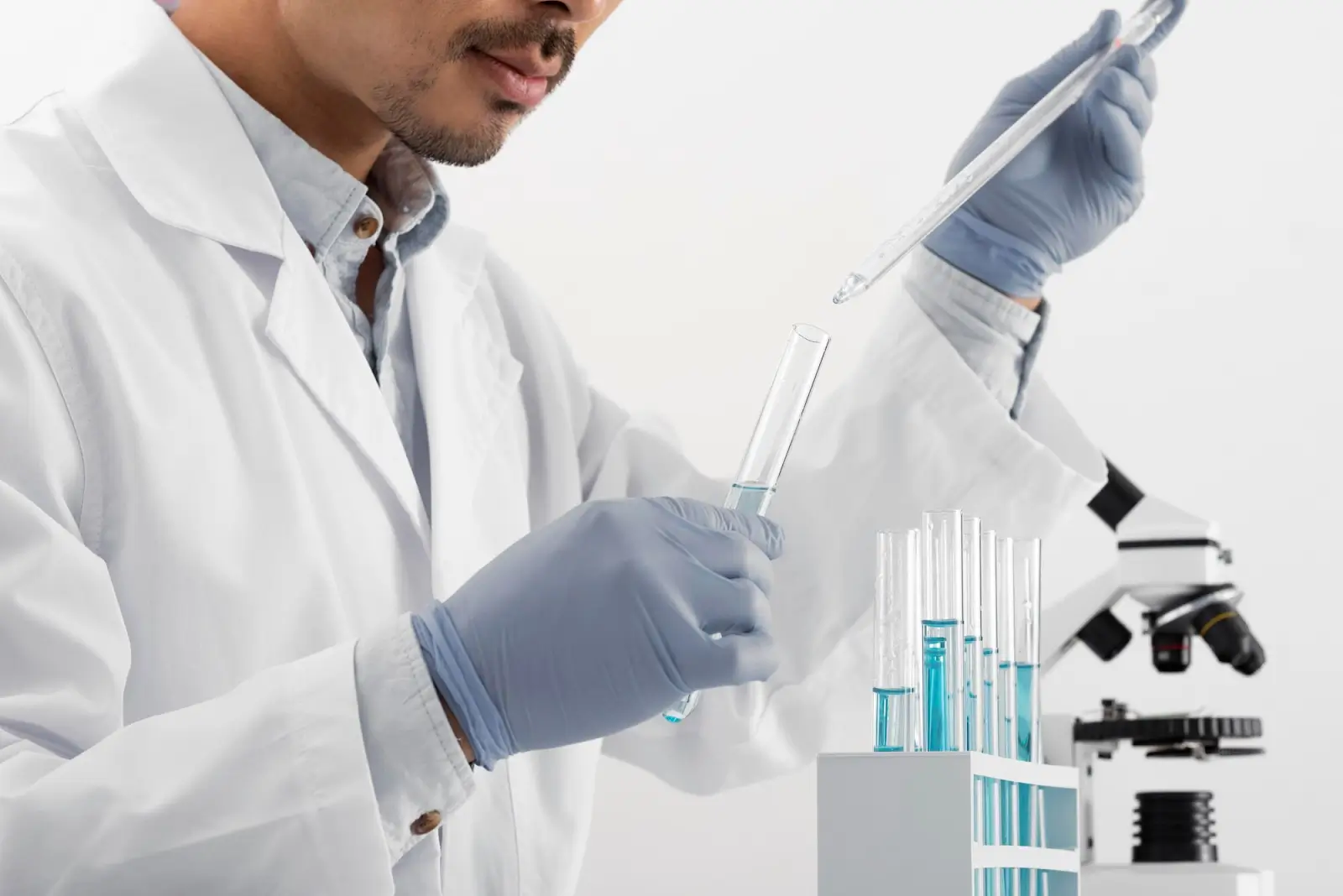 A scientist wearing a lab coat and gloves uses a pipette to transfer liquid into a test tube, with more test tubes in a rack and a microscope in the background.