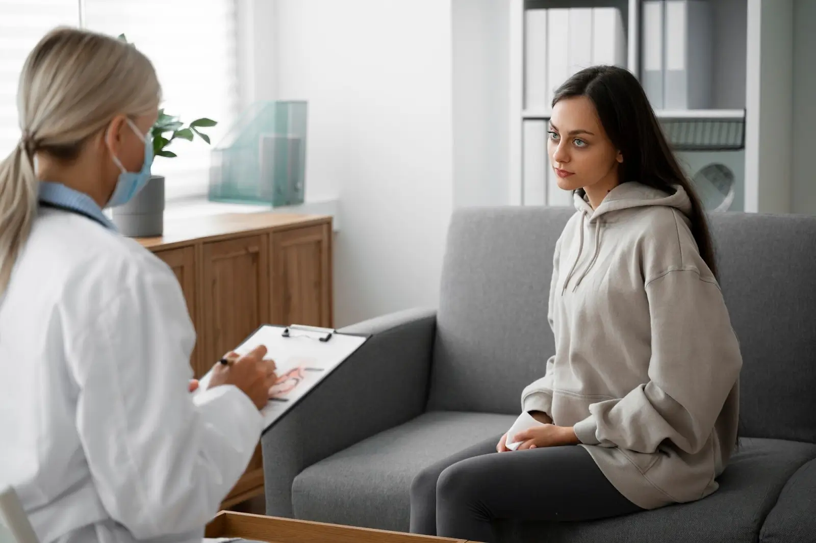 A young woman in a hoodie sits on a gray couch, attentively listening to a healthcare professional in a white coat taking notes on a clipboard in a bright office setting.