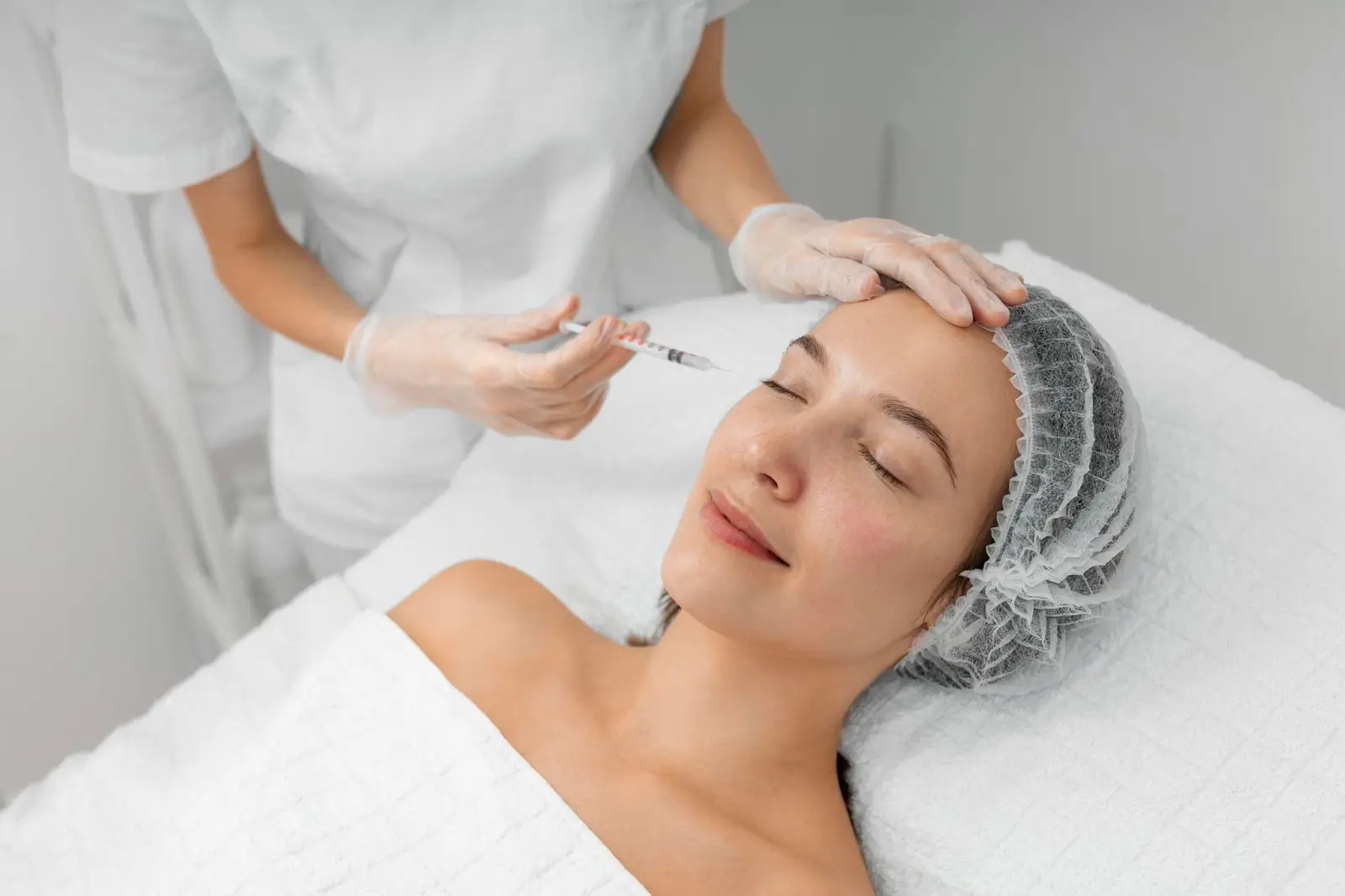 A woman lying on a treatment bed with her eyes closed receives an injection in her face from a medical professional wearing gloves and a white uniform. The woman wears a hair cap and appears calm and relaxed.