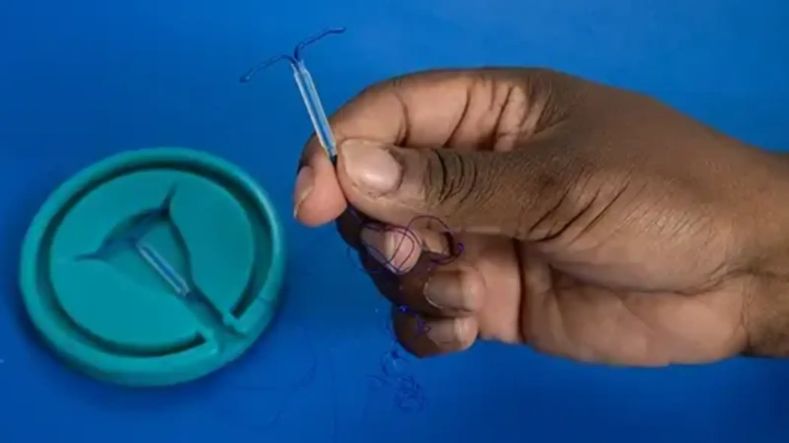 A hand holding an intrauterine device (IUD) above a blue surface, with another IUD placed in a round green container nearby.