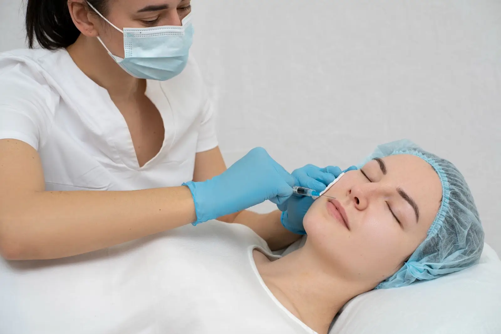 A healthcare professional wearing a mask and gloves injects a syringe into the cheek of a woman lying down with a hair cover, likely during a cosmetic or medical procedure.