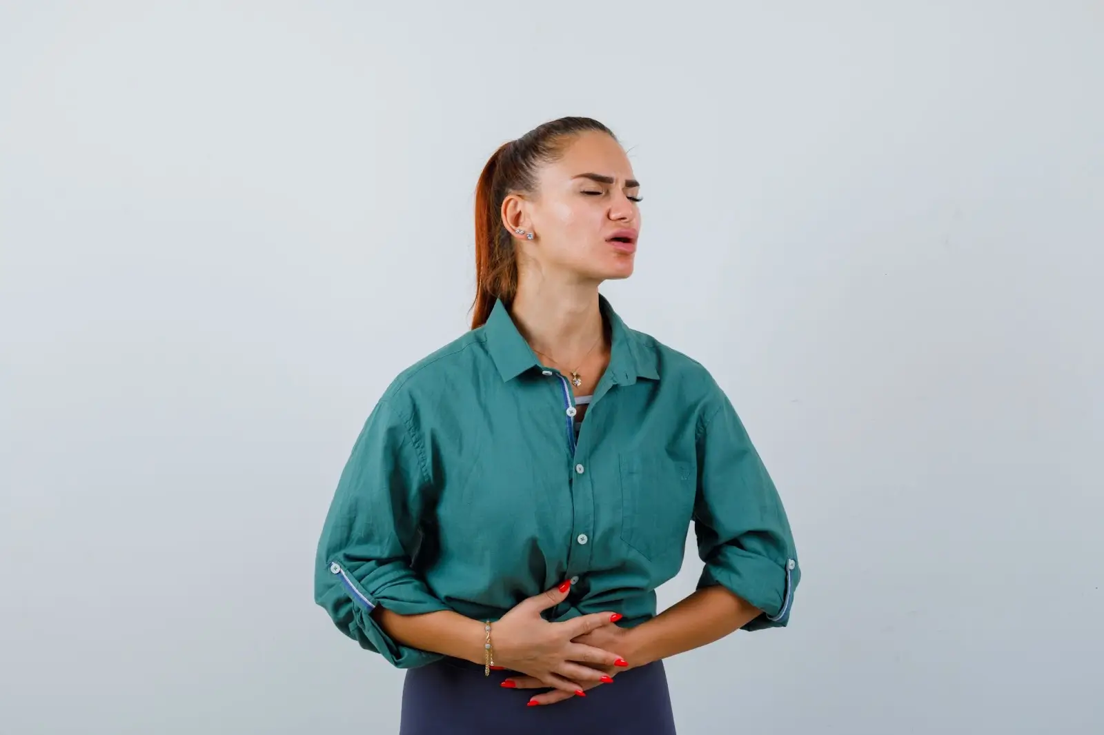 A woman wearing a green shirt stands against a plain background, holding her stomach with both hands and appearing to be in pain or discomfort, with her eyes closed and brows furrowed.