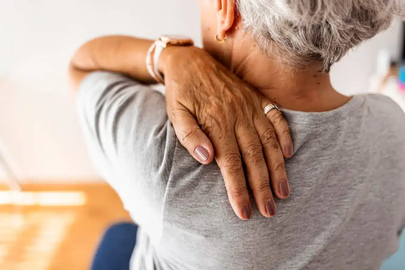An older adult with short gray hair wearing a gray shirt touches their shoulder, suggesting pain or discomfort. The image is taken from behind, highlighting the hand resting on the upper back.
