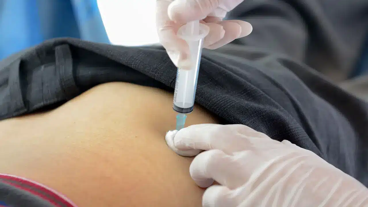 A close-up of a medical professional wearing gloves administering an injection with a syringe into a person's lower back or hip area. The person is lying down and their shirt is lifted to expose the injection site.