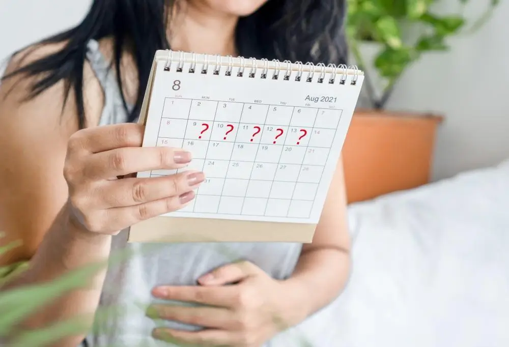 A person holds an August 2021 calendar with question marks on several dates, appearing concerned while touching their stomach; indoor setting with plants in the background.