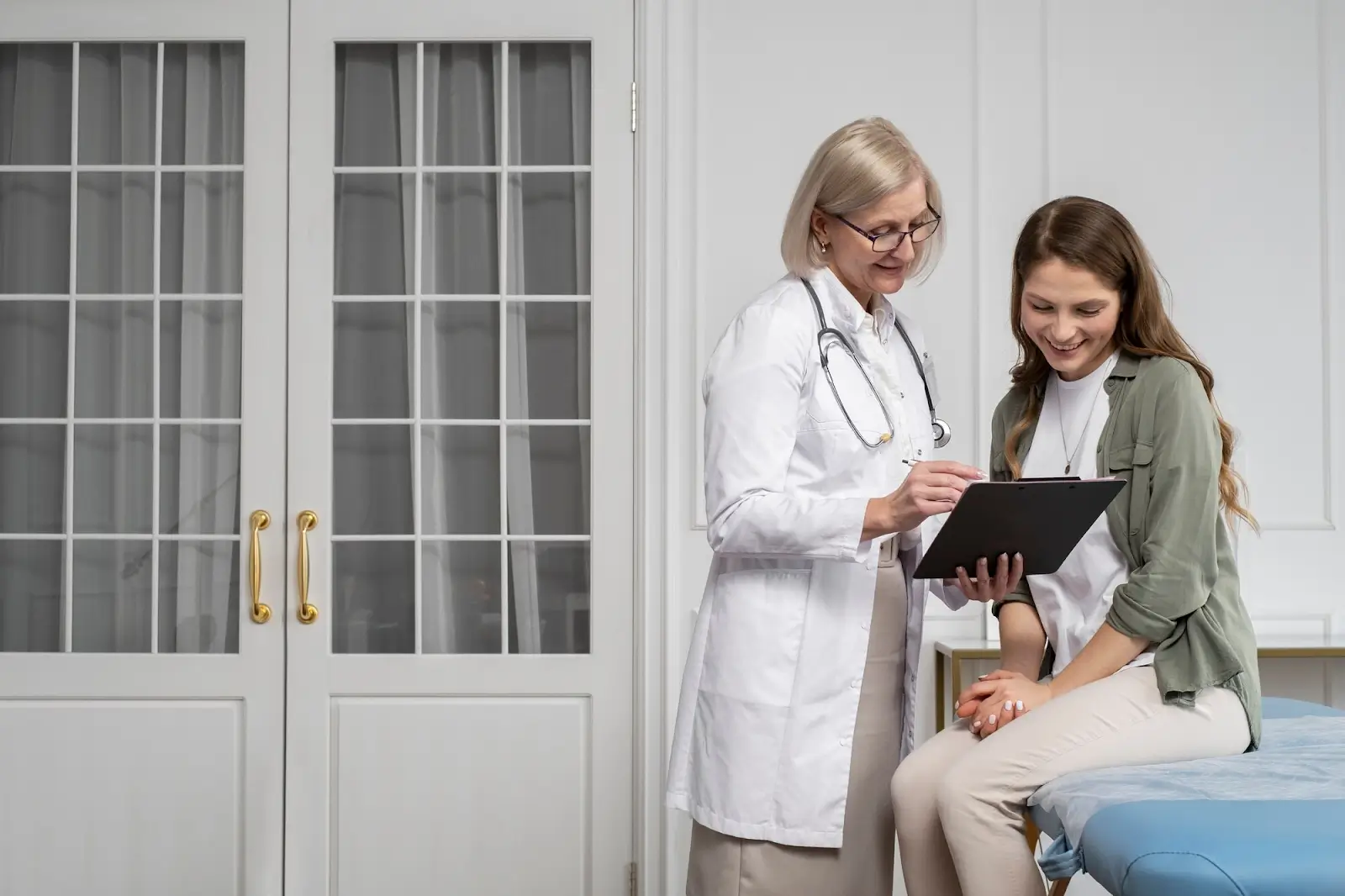 A doctor in a white coat shows a clipboard to a smiling woman sitting on an exam table in a medical office with glass doors in the background.
