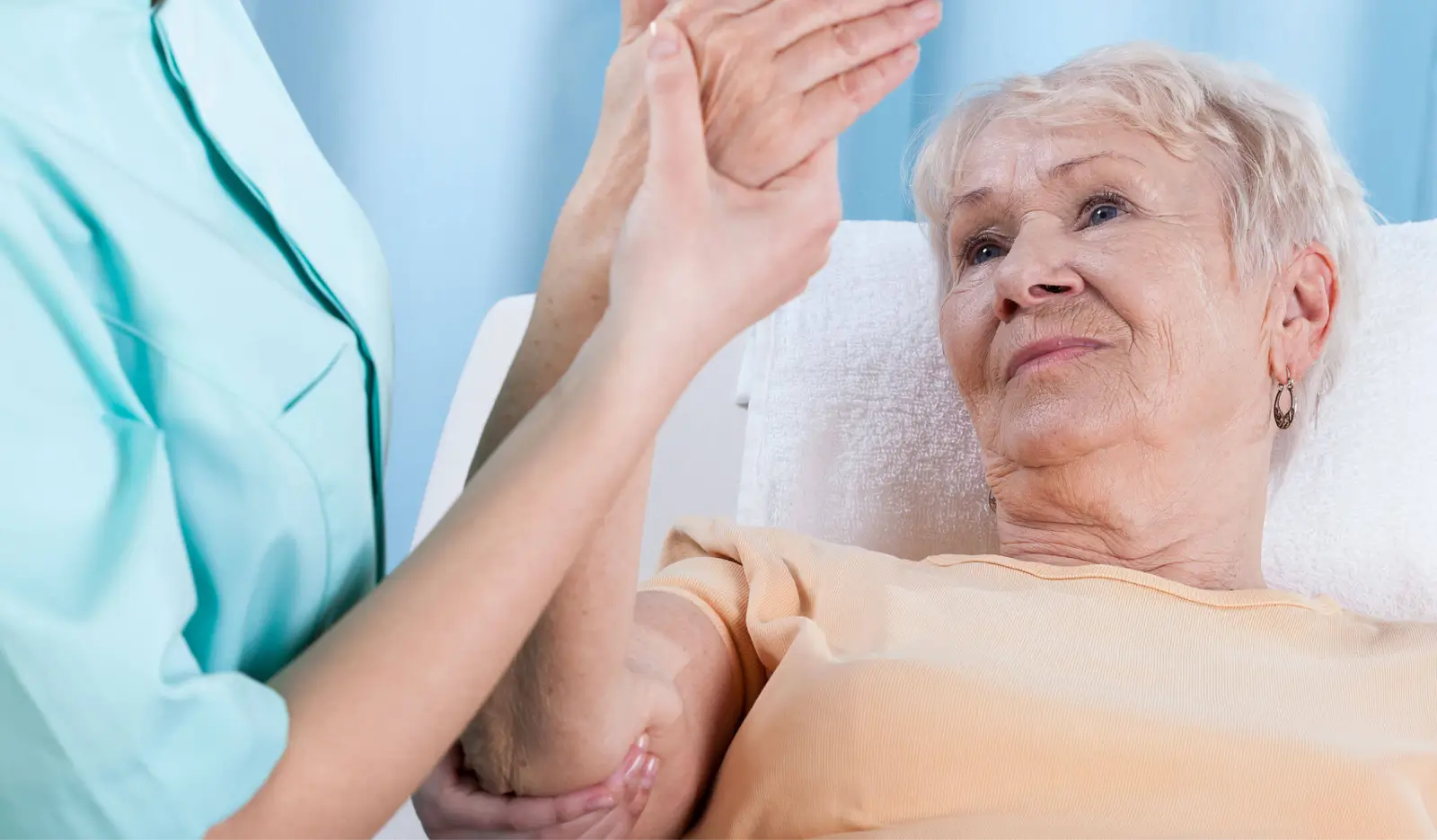 A nurse or caregiver in a blue uniform helps an elderly woman with gray hair perform an arm exercise while the woman lies on a bed, looking up at the caregiver.