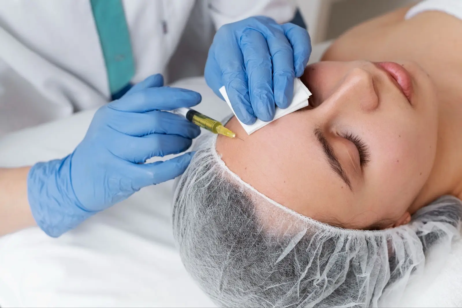 A person wearing a hair net lies with eyes closed while a medical professional in blue gloves injects a syringe into their forehead, possibly administering a cosmetic treatment.