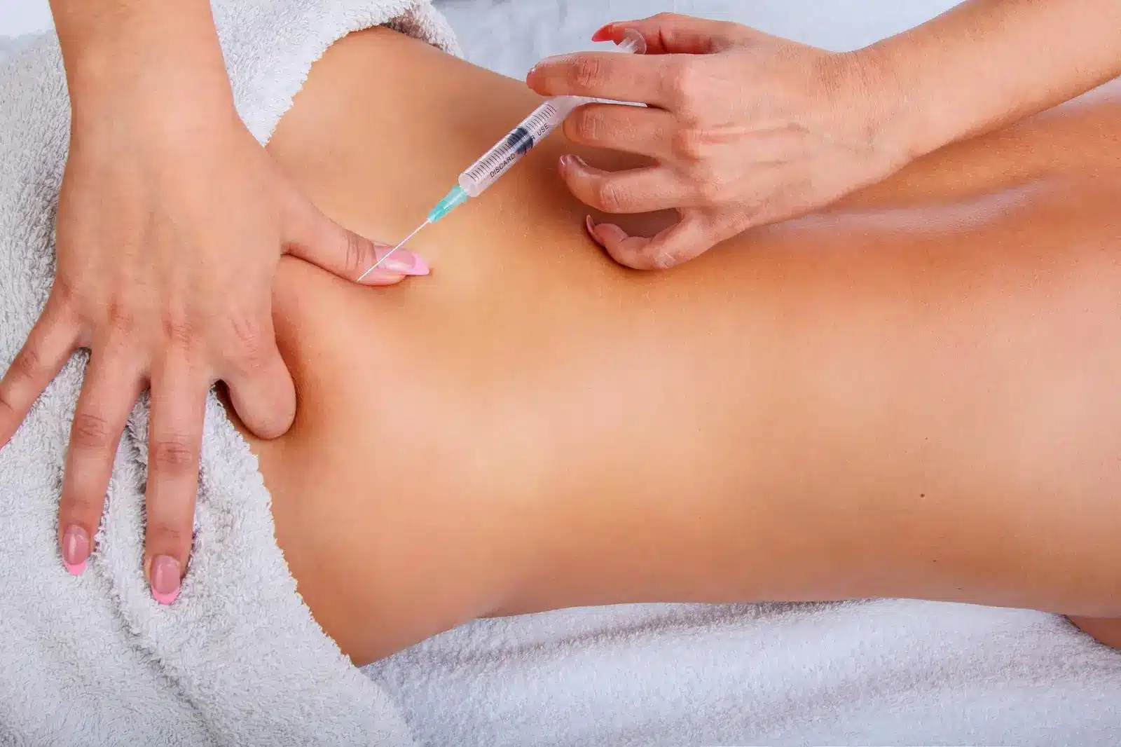 A person lying face down with a towel covering their lower body while another person injects a syringe into their lower back.
