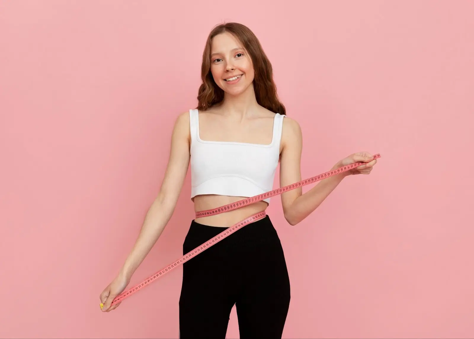 A smiling young woman in a white crop top and black pants wraps a measuring tape around her waist while standing against a pink background.