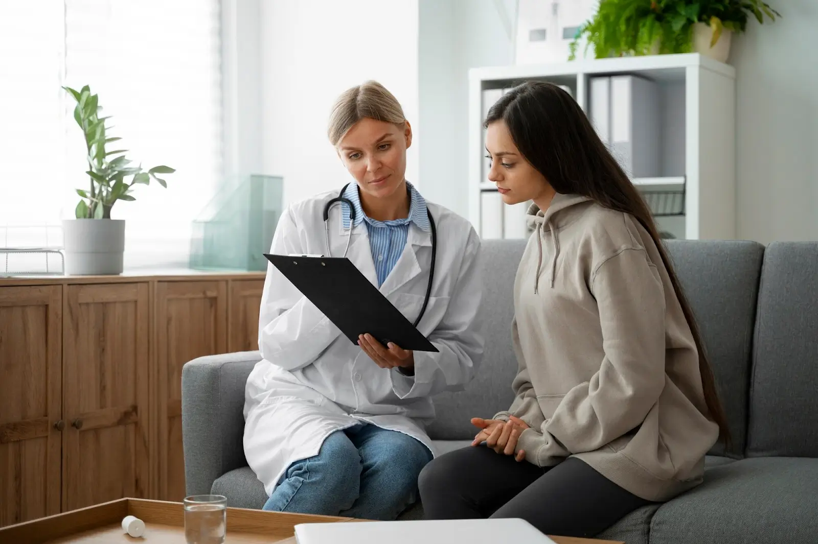A doctor in a white coat shows a clipboard to a young woman sitting next to her on a couch in a bright office, discussing medical information together.