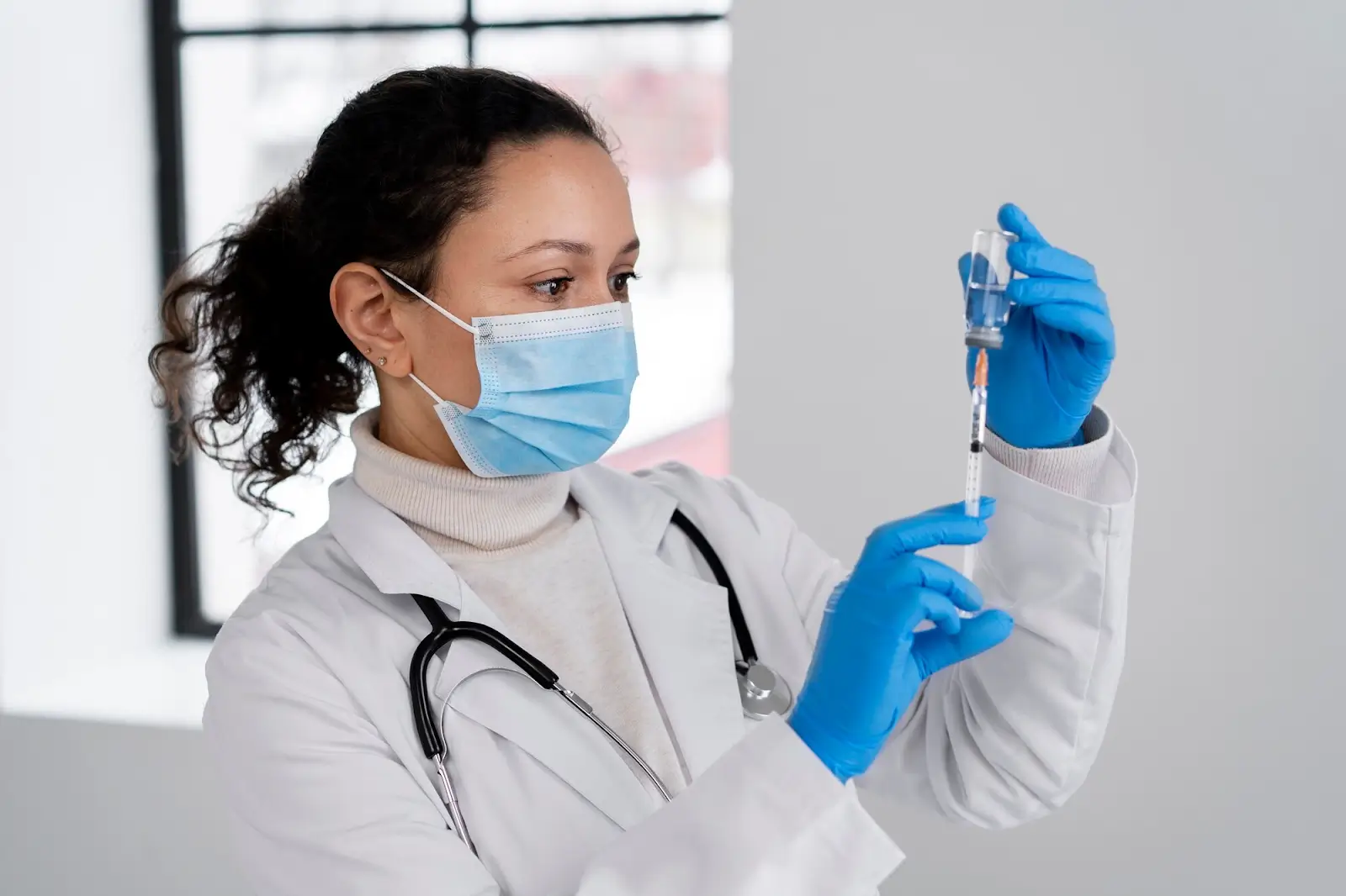 A female doctor wearing a white coat, blue gloves, and a face mask is preparing a syringe with liquid from a vial. She is indoors with a stethoscope around her neck.