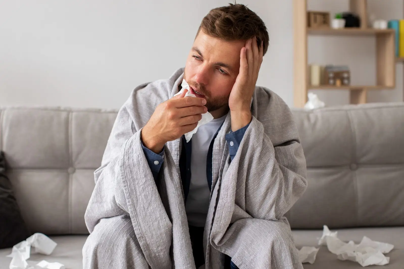 A man sits on a couch wrapped in a blanket, holding a tissue to his nose. He looks unwell and tired, with several crumpled tissues around him, suggesting he may have a cold or flu.