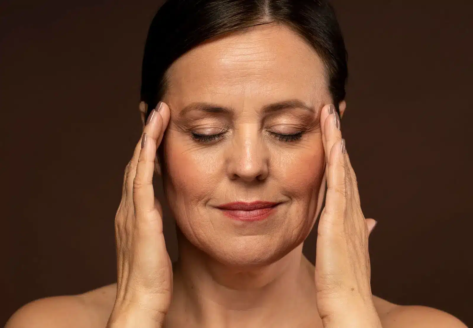 A woman with closed eyes gently touches her temples with both hands. She has a calm, relaxed expression and a neutral brown background.