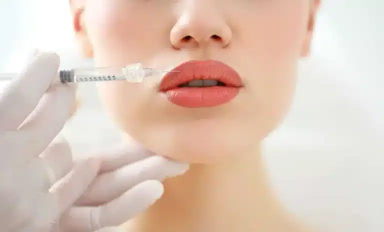 Close-up of a person's face receiving a lip injection. A gloved hand is holding a syringe near their lips, which are slightly parted. The background is blurred, emphasizing the focus on the lips and the procedure.
