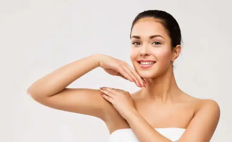 A smiling woman with dark hair pulled back is posing against a light gray background. She has one hand resting on her shoulder and the other hand gracefully positioned near her chin. She is wearing a white strapless top.