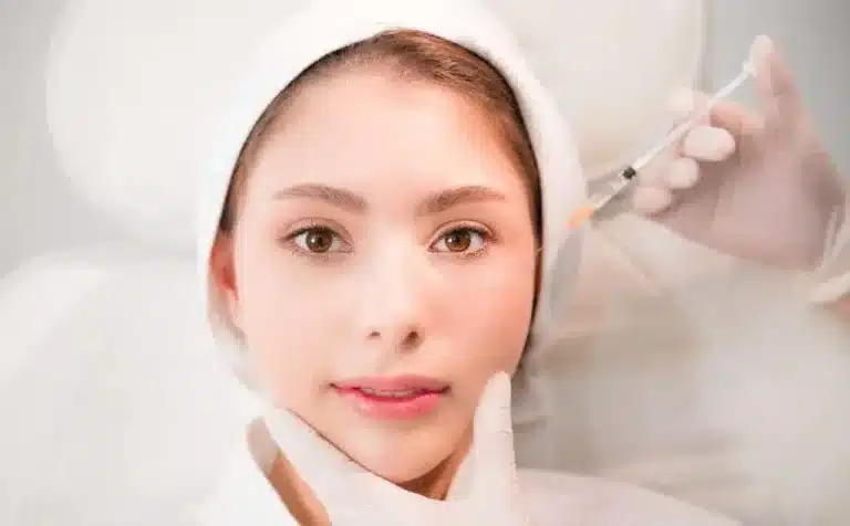 A person with light skin, wearing a white headband, is receiving an injection on their cheek. There is a gloved hand gently touching their chin, and another gloved hand holding a syringe near their face. The background is bright and blurred.