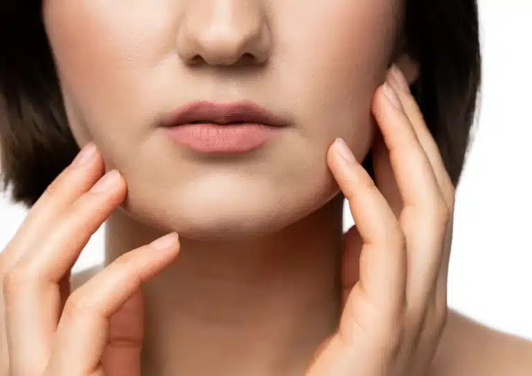 Close-up image of the lower face of a woman with smooth skin, light pink lips, and short, dark hair. Her hands are gently touching her jawline and cheeks, emphasizing a skincare routine, possibly featuring Aqualyx treatment. The background is white and uncluttered.