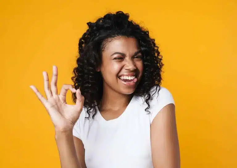 A person with curly hair wearing a white t-shirt is standing against a vibrant yellow background. They are smiling, winking, and making an "OK" gesture with their right hand, as if giving a nod of approval to the results of their latest Aqualyx treatment.