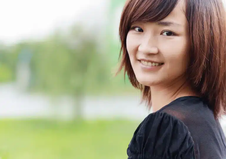 A person with short brown hair smiles at the camera. They are wearing a black top and stand in front of a blurred outdoor background with greenery.